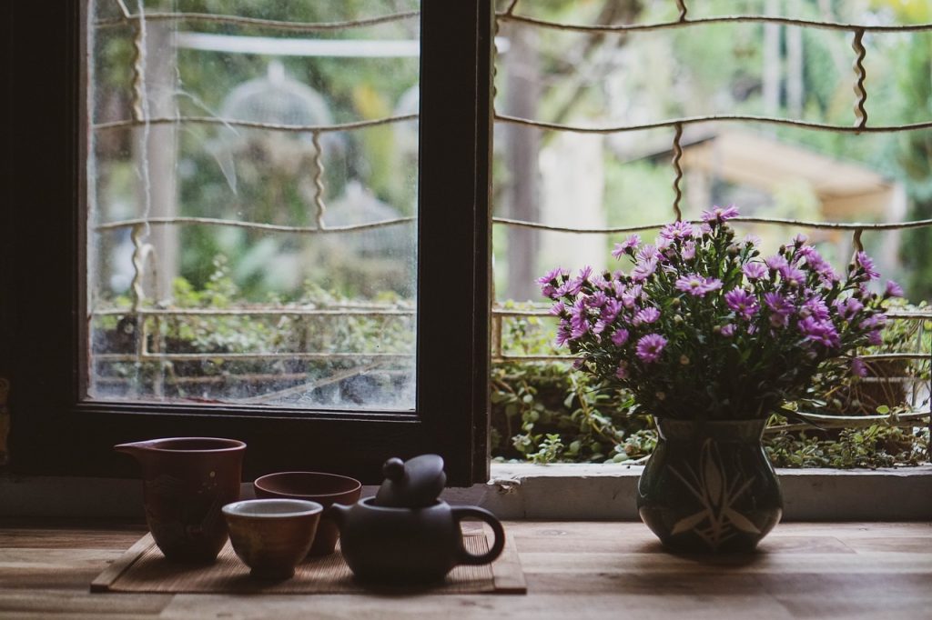 A Japanese-style tea set sits next to a pot of purple flowers next to a large window, perfect for self-care under the full moon.